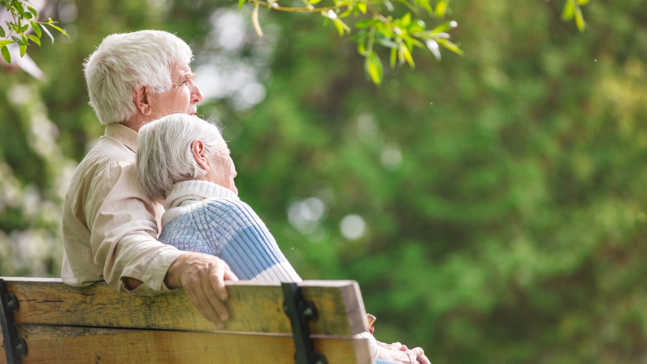 An older couple sitting close together on a park bench under green trees